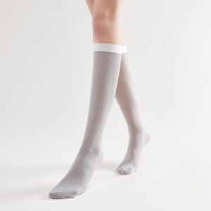May include: A pair of knee-high socks in a light gray color with a white band at the top. The socks have a subtle shimmer and are worn by a person. The background is a plain white.