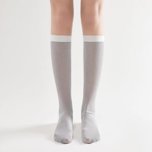May include: A pair of knee-high socks in a light gray color with a white band at the top. The socks are made of a thin, sheer material. The image is a close-up of the socks, showing the details of the fabric and the fit.