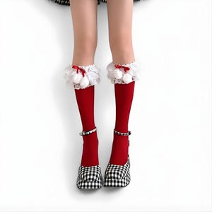 May include: Red knee-high socks with white lace trim, small white pom-poms, and red bows. The socks are worn with black and white checked Mary Jane shoes with ankle straps. The background is white.