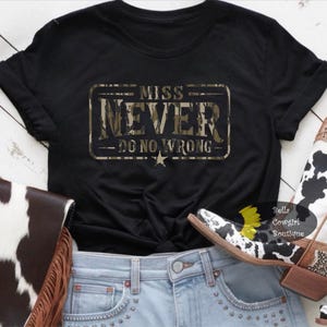 May include: Black t-shirt with the text "MISS NEVER DO NO WRONG" in a camouflage design. The shirt is paired with light blue jeans, a cowhide purse, and cow print boots.