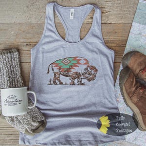 May include: Gray tank top with a graphic of a bison with a Southwestern design on its back. The tank top is on a wooden surface with a mug, socks, boots, and a map.