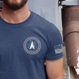 May include: A navy blue t-shirt with a white graphic of the United States Space Force logo. The logo features a rocket ship and the text "United States Space Force Department of the Air Force". The shirt also has a small American flag patch on the right sleeve.