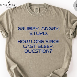 May include: Khaki-colored t-shirt with the text "GRUMPY. ANGRY. STUPID. HOW LONG SINCE LAST SLEEP, QUESTION?" in blue. The shirt has short sleeves and a crew neck.