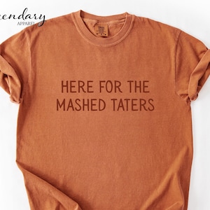 May include: A rust-colored t-shirt with the text "HERE FOR THE MASHED TATERS" printed in a darker shade. The shirt has a crew neck and short sleeves, with the sleeves rolled up.
