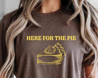 May include: Brown t-shirt with the text "HERE FOR THE PIE" in yellow above a yellow line drawing of a slice of pie with whipped cream. The shirt is a casual style.