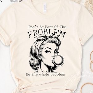 May include: Cream-colored t-shirt with the text "Don't Be Part Of The PROBLEM, Be the whole problem." A black and white illustration of a woman blowing a bubble gum bubble is in the center. The shirt has rolled-up sleeves.