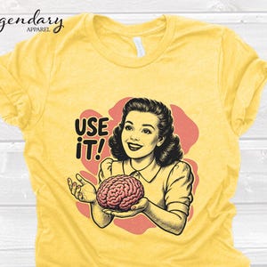 Pu&ograve; includere: T-shirt giallo con un'illustrazione retr&ograve; di una donna che tiene in mano un cervello. Il testo "USE IT!" &egrave; in grassetto. La maglietta ha un girocollo e maniche corte.