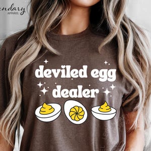 May include: Brown t-shirt with the text "deviled egg dealer" in white, along with illustrations of deviled eggs. The shirt features a crew neck and short sleeves. The Legendary Apparel logo is visible.