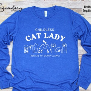 May include: A long-sleeved, heather royal blue t-shirt with the text "Childless Cat Lady" in white. The text is surrounded by a row of white cat paw prints and a heart. The text "(Beware of Sharp Claws)" is printed below the cat paw prints.
