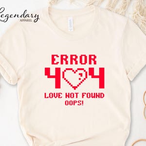 May include: Cream-colored t-shirt with the text "ERROR 404 LOVE NOT FOUND OOPS!" in red pixelated font. The design includes a pixelated heart symbol. The shirt has short sleeves and a crew neck.