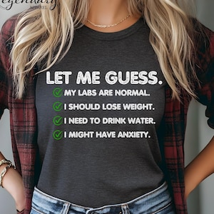 May include: A dark gray t-shirt with a white graphic that says "Let me guess." and lists a series of checkboxes with humorous statements, including "My labs are normal", "I should lose weight", "I need to drink water", and "I might have anxiety".