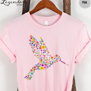 May include: Pink t-shirt with a hummingbird design made of colorful flowers. The shirt is a light pink color and has a crew neck.