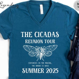 May include: A teal V-neck t-shirt with white text that reads "THE CICADAS REUNION TOUR" and "SUMMER 2025". A detailed white illustration of a cicada is centered below the text. The shirt also includes the text "CENTURIES IN THE MAKING, THE BROOD IS BACK."