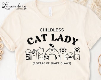 May include: A white t-shirt with the text "Childless Cat Lady" in black lettering. The text is surrounded by a line of black cat paw prints and a heart. The text "(Beware of Sharp Claws)" is printed below the paw prints.