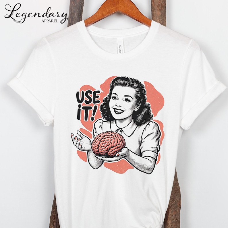 Pu&ograve; includere: T-shirt bianca con un'illustrazione retr&ograve; di una donna che tiene in mano un cervello. Il design include il testo "USE IT!" in grassetto. La donna sorride e il cervello &egrave; rosa.