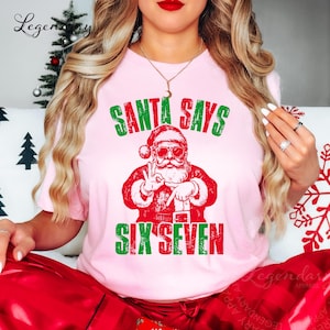 May include: A light pink t-shirt with a graphic of Santa Claus wearing sunglasses and making a hand gesture. The text "SANTA SAYS" is above the Santa graphic, and "SIX SEVEN" is below, all in green and red.