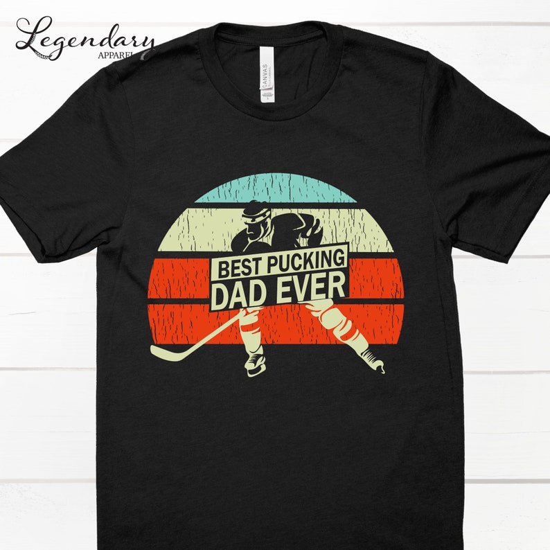 May include: Black t-shirt with a retro sunset graphic featuring a hockey player holding a sign that says "Best Pucking Dad Ever".