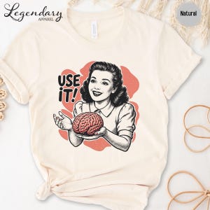 Pu&ograve; includere: T-shirt color crema con grafica retr&ograve; di una donna che tiene in mano un cervello, con la scritta "USE IT!". Il design presenta uno stile vintage con sfondo rosso. La maglietta ha maniche corte e scollo tondo.