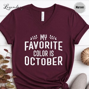 A burgundy-colored t-shirt with the text "My favorite color is October" printed on it, along with decorative leaves. The t-shirt is displayed on a white background, and there are some decorative elements such as a plant and a ball visible in the background.