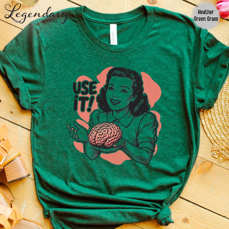 Pu&ograve; includere: T-shirt verde erica con un'illustrazione retr&ograve; di una donna che tiene in mano un cervello, con la scritta "USE IT!". Il design &egrave; nei toni del rosa, verde e nero. La maglietta &egrave; realizzata in materiale morbido.