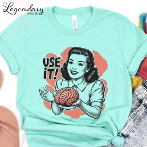 Pu&ograve; includere: T-shirt verde acqua chiaro con una grafica vintage di una donna che tiene in mano un cervello e la scritta "USE IT!". La maglietta &egrave; realizzata in materiale morbido, ideale per l'uso quotidiano. Il design &egrave; stampato sul davanti.