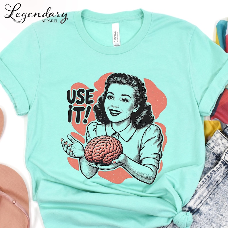 Pu&ograve; includere: T-shirt verde acqua chiaro con una grafica vintage di una donna che tiene in mano un cervello e la scritta "USE IT!". La maglietta &egrave; realizzata in materiale morbido, ideale per l'uso quotidiano. Il design &egrave; stampato sul davanti.