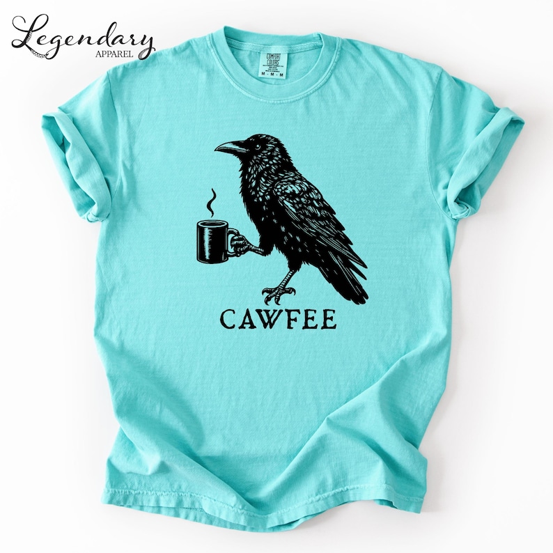 A turquoise t-shirt with a black illustration of a crow holding a coffee cup and the text "CAWFEE" below it.