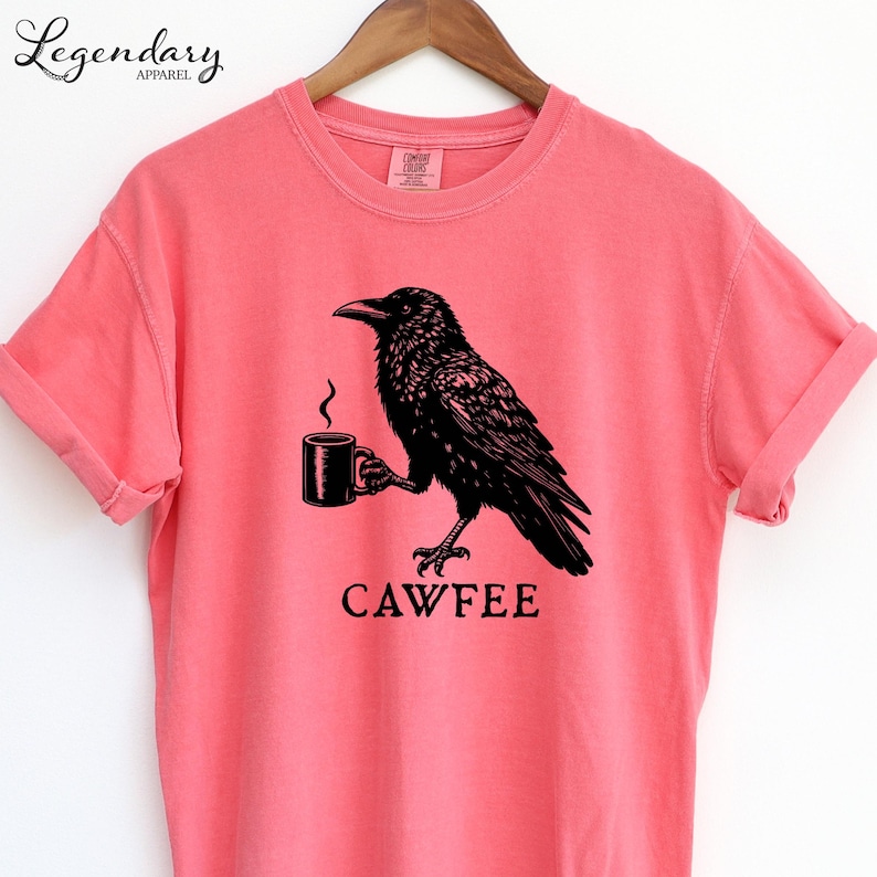 A pink t-shirt with a black illustration of a crow holding a coffee cup and the text "CAWFEE" below it.