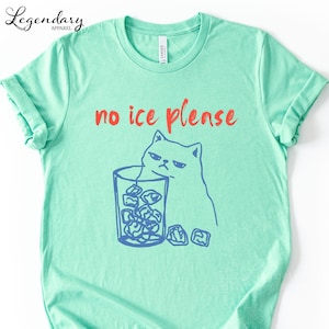 May include: A light green t-shirt with the text "no ice please" in red. A blue illustration of a grumpy cat is next to a glass of ice cubes. The shirt has rolled-up sleeves.