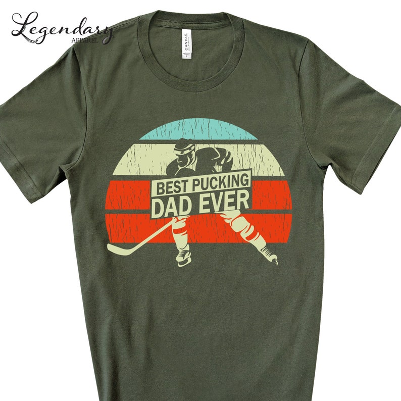 May include: Green t-shirt with a retro sunset graphic featuring a hockey player and the text "Best Pucking Dad Ever".