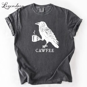The image is a gray t-shirt with a white graphic design depicting a bird holding a cup of coffee. The text "CAWFEE" is printed below the bird and cup.