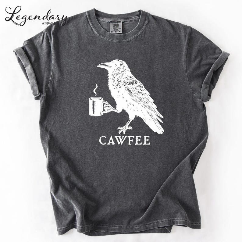 The image is a gray t-shirt with a white graphic design depicting a bird holding a cup of coffee. The text "CAWFEE" is printed below the bird and cup.
