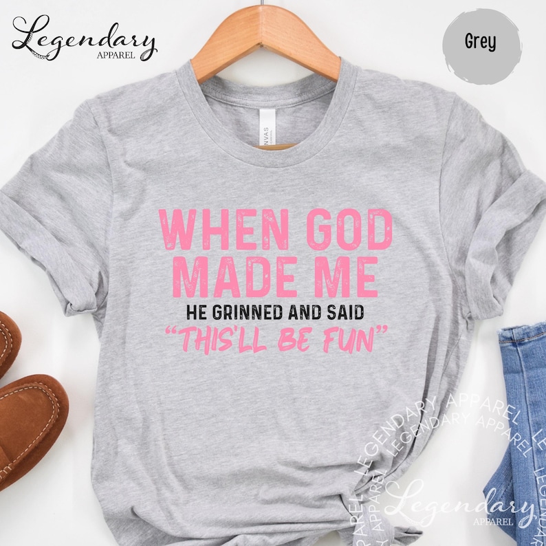 A grey t-shirt with a humorous text printed on it, hanging on a wooden hanger.