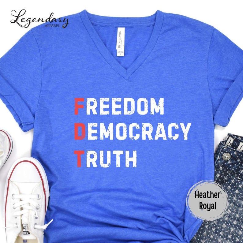 Op de afbeelding: Heather Royal blauw V-hals T-shirt met de woorden "FREEDOM DEMOCRACY TRUTH" in wit en rood gedrukt. Het shirt heeft een label met "Bella + Canvas". Witte sneakers en een blauwe jeans zijn zichtbaar in de afbeelding.