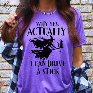 Może przedstawiać: Fioletowy t-shirt z krótkim rękawem z czarną grafiką przedstawiającą czarownicę na miotle i kota. Napis głosi: WHY YES, ACTUALLY I CAN DRIVE A STICK. Koszulka ma okrągły dekolt i jest noszona z czarno-białą koszulą w kratę.