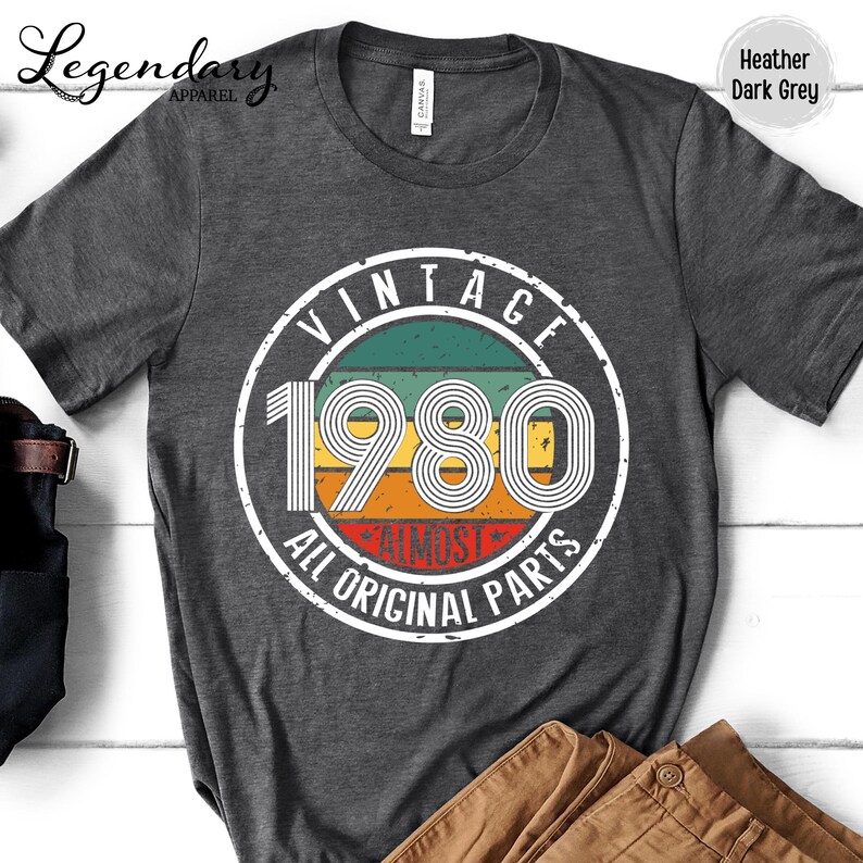 May include: Heather dark grey t-shirt with a circular graphic. The graphic reads "VINTAGE 1980 ALL ORIGINAL PARTS" in white lettering, with a retro color scheme of green, yellow, orange, and red. The shirt has a crew neck and short sleeves.