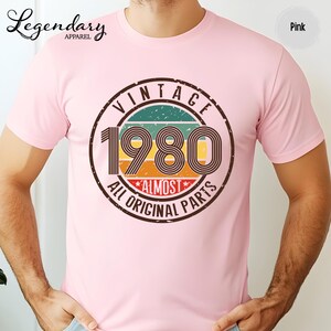May include: Pink t-shirt with a vintage 1980 graphic. The graphic is a circular design with the words "VINTAGE 1980 ALL ORIGINAL PARTS" in a retro font. The shirt also has the "Legendary Apparel" logo.