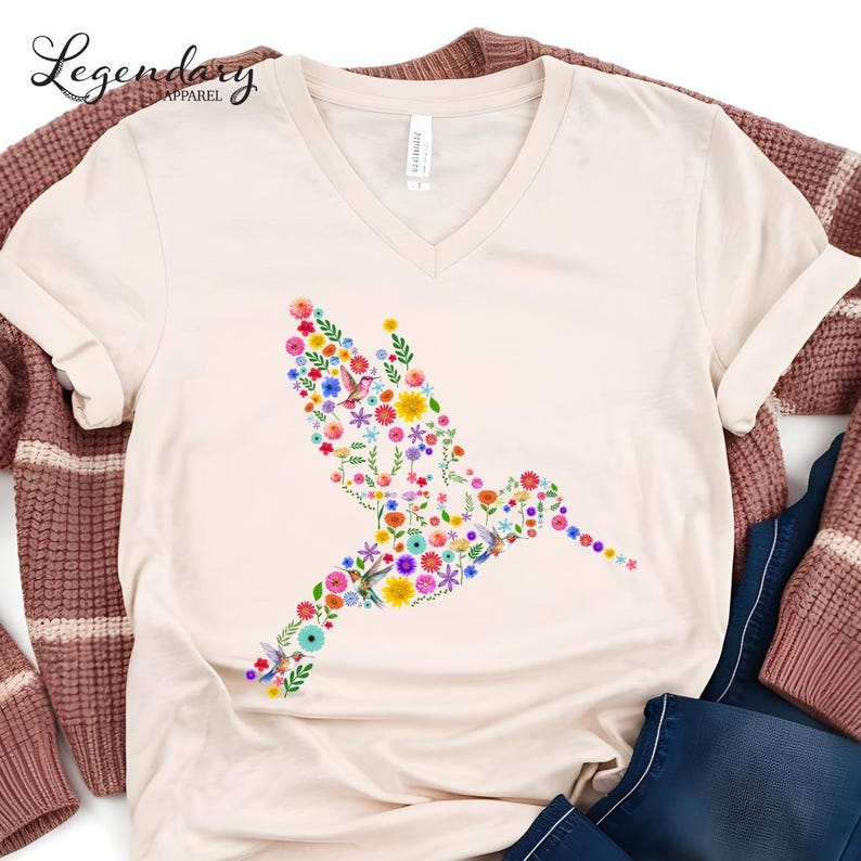 May include: A beige v-neck t-shirt with a floral hummingbird design. The hummingbird is made up of colorful flowers and leaves.