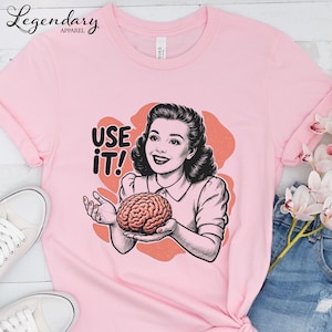 Pu&ograve; includere: T-shirt rosa chiaro con grafica retr&ograve; di una donna che tiene in mano un cervello, con la scritta "USE IT!". Il design &egrave; in nero, bianco e corallo. La maglietta ha maniche arrotolate e girocollo.