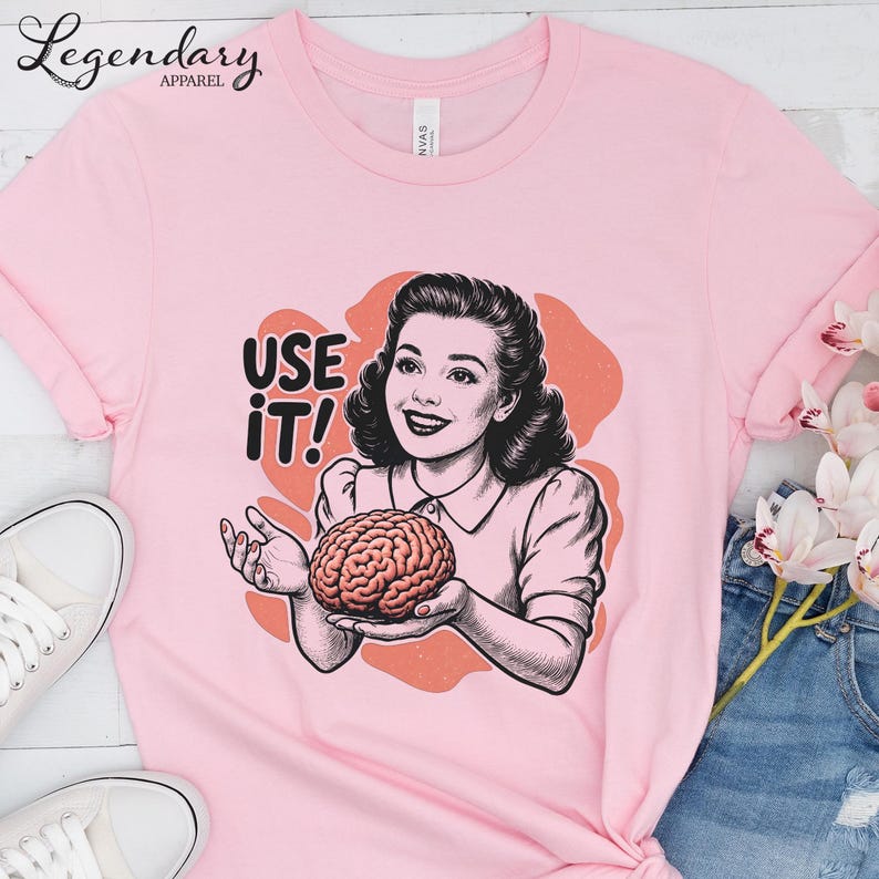 Pu&ograve; includere: T-shirt rosa chiaro con grafica retr&ograve; di una donna che tiene in mano un cervello, con la scritta "USE IT!". Il design &egrave; in nero, bianco e corallo. La maglietta ha maniche arrotolate e girocollo.