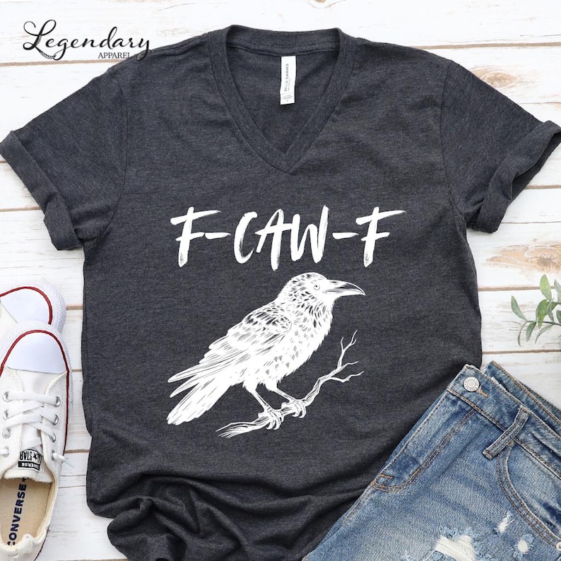 Pode incluir: T-shirt cinza escuro com decote em V com um gr&aacute;fico branco de um corvo empoleirado em um galho e o texto "F-CAW-F" acima. A imagem inclui jeans azul e t&ecirc;nis brancos.