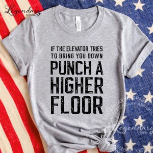 May include: A heather gray t-shirt with the text "IF THE ELEVATOR TRIES TO BRING YOU DOWN PUNCH A HIGHER FLOOR" in black block letters. The shirt is displayed against a backdrop of an American flag.