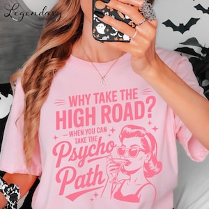 May include: Pink t-shirt with the text "WHY TAKE THE HIGH ROAD? WHEN YOU CAN TAKE THE Psycho Path" in a retro font. The graphic includes a woman drinking a cocktail. The shirt is a light pink color.