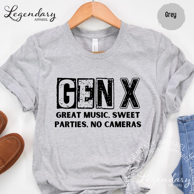 A grey t-shirt with the text "GENX" printed on it, along with the text "GREAT MUSIC, SWEET PARTIES. NO CAMERAS" below it. The t-shirt is hanging on a wooden hanger, and there are some brown shoes visible in the background.