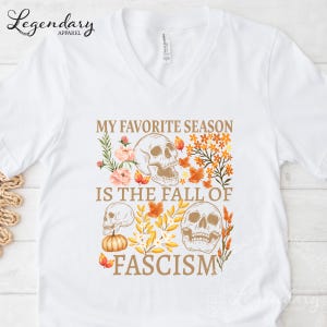 May include: White v-neck t-shirt with a graphic design featuring skulls, autumn leaves, and the text "MY FAVORITE SEASON IS THE FALL OF FASCISM". The design includes a pumpkin and floral elements.
