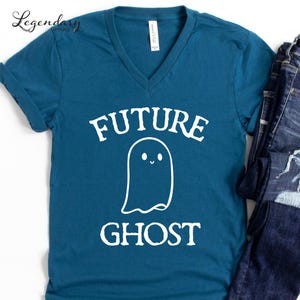 May include: Teal v-neck t-shirt with the words "FUTURE GHOST" in white, along with a cute ghost graphic. The shirt is paired with blue jeans. The shirt is made of a soft material.