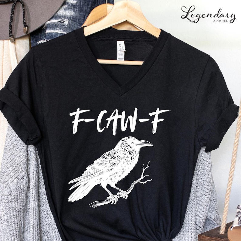 Pode incluir: Camiseta preta com decote em V com um desenho branco de um corvo num galho e o texto "F-CAW-F". A camiseta est&aacute; pendurada num cabide de madeira.
