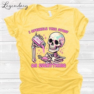 May include: Yellow t-shirt with a graphic of a skeleton sprinkling pink "sarcasm" on everything. The skeleton has a colorful skull and is holding a jar labeled "sarcasm". The text "I sprinkle this stuff on everything" is also on the shirt.