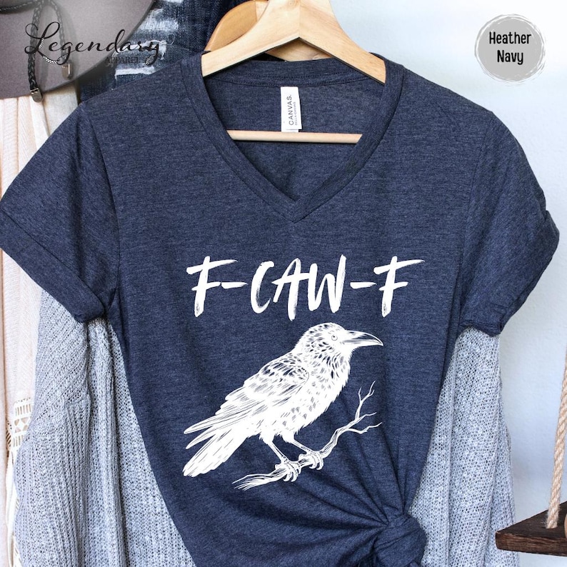 Pode incluir: Camiseta com decote em V azul marinho mesclado com o texto "F-CAW-F" e uma ilustra&ccedil;&atilde;o branca de um corvo em um galho. A camiseta est&aacute; pendurada em um cabide de madeira.