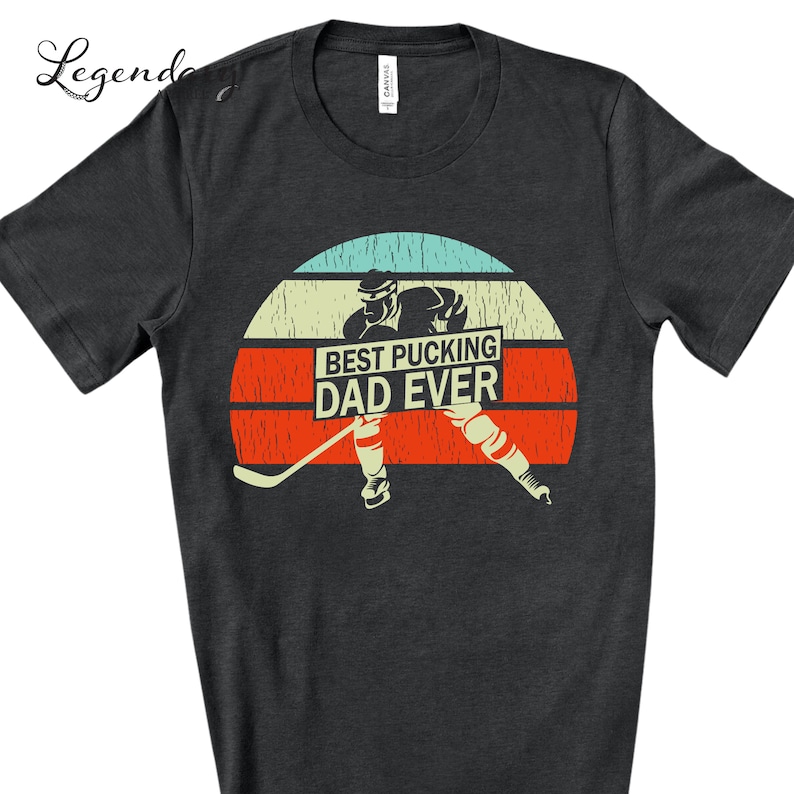 May include: A dark gray t-shirt with a vintage-style graphic of a hockey player skating with a hockey stick. The graphic includes a sunset background and the text "Best Pucking Dad Ever".
